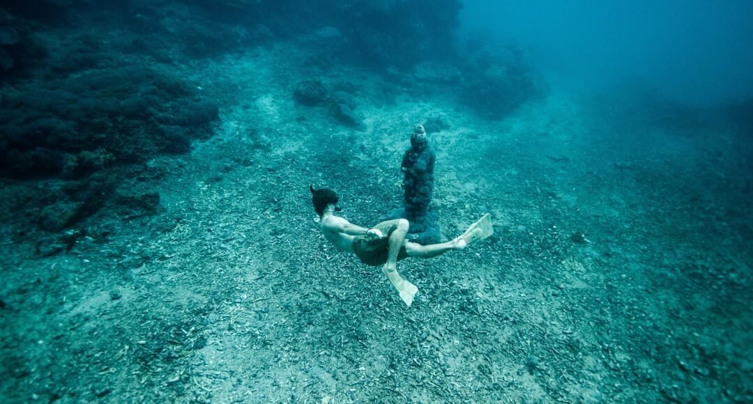 Persona buceando cerca de una estatua sumergida en un fondo marino con corales y agua clara.