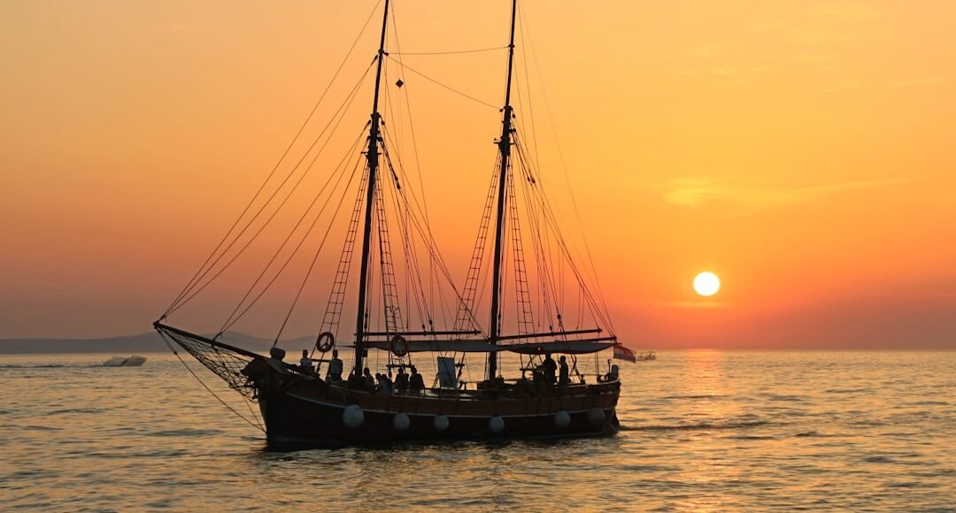 Velero navegando al atardecer con el sol poniéndose en el horizonte y el cielo teñido de tonos naranjas.