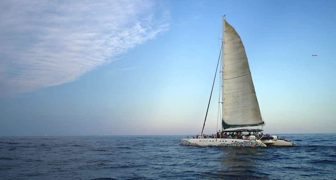 Catamarán a vela navegando en mar abierto bajo un cielo despejado con un avión visible a lo lejos.